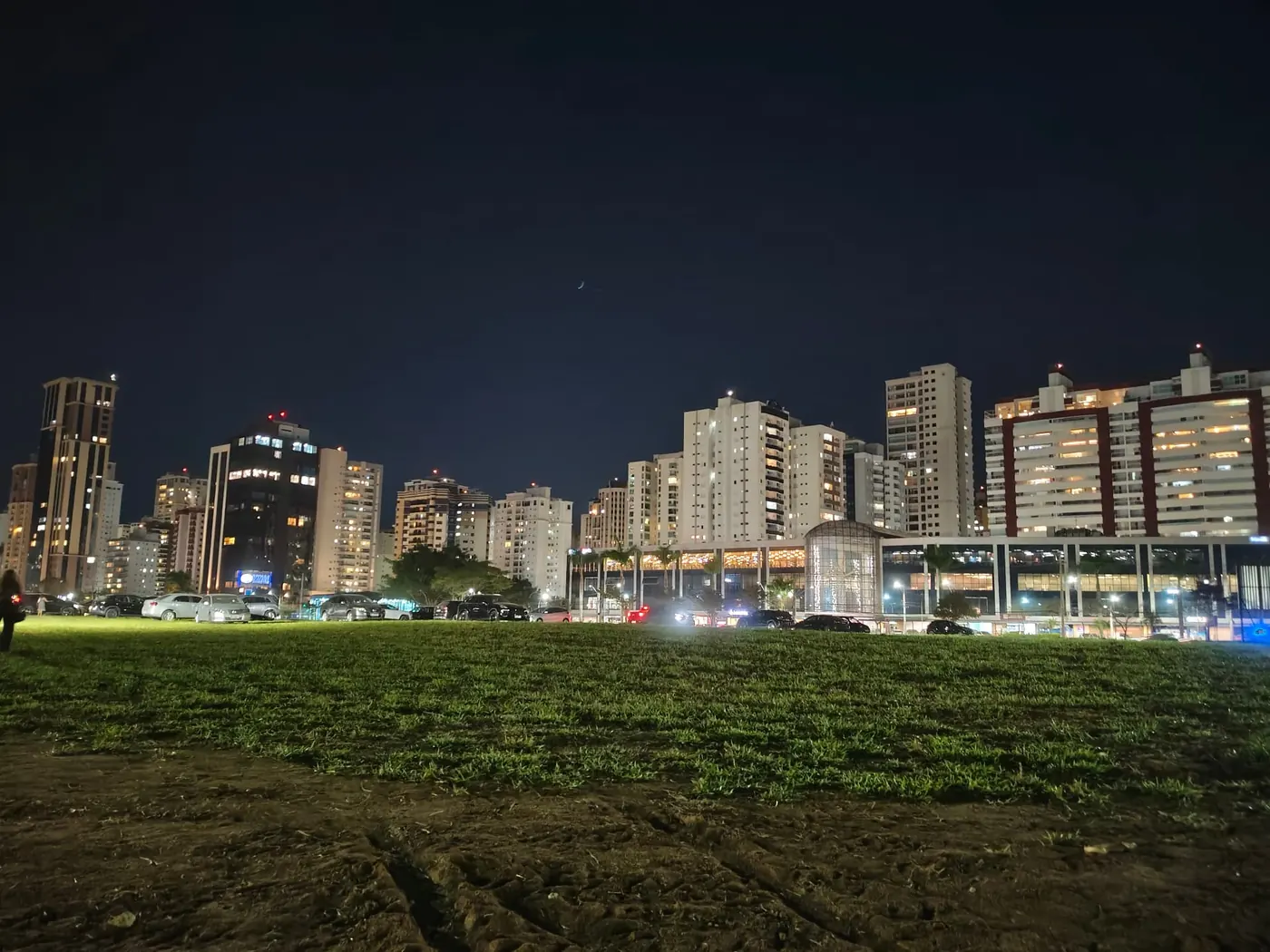 Vista noturna do terreno gramado do Parque Una com o skyline do Jardim Aquarius ao fundo
